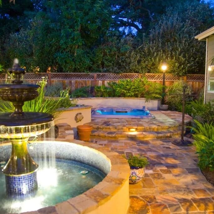 Beautifully crafted backyard fountain with custom stonework, surrounded by lush greenery and atmospheric lighting in Los Altos, CA. A tranquil outdoor oasis.